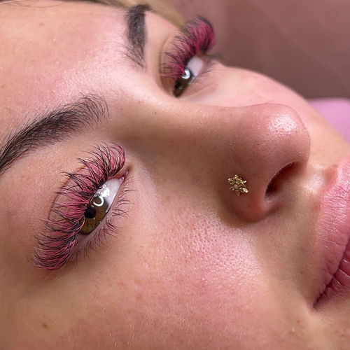 A woman laying down, looking into the camera as she shows off her new pink colored lash extensions.
