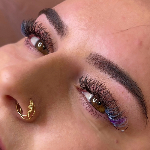A woman laying down, looking into the camera as she shows off her new blue & purple colored lash extensions.